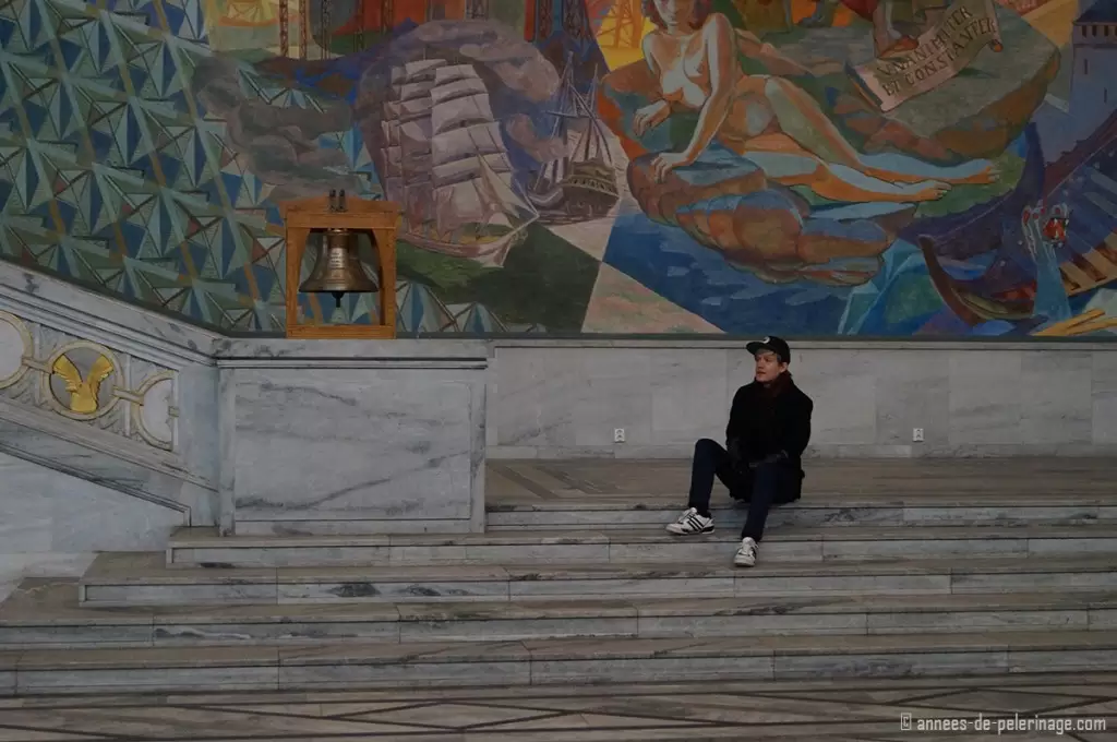 Me sitting inside the city hall in oslo on some raised stairs with a very colorful mural in the background