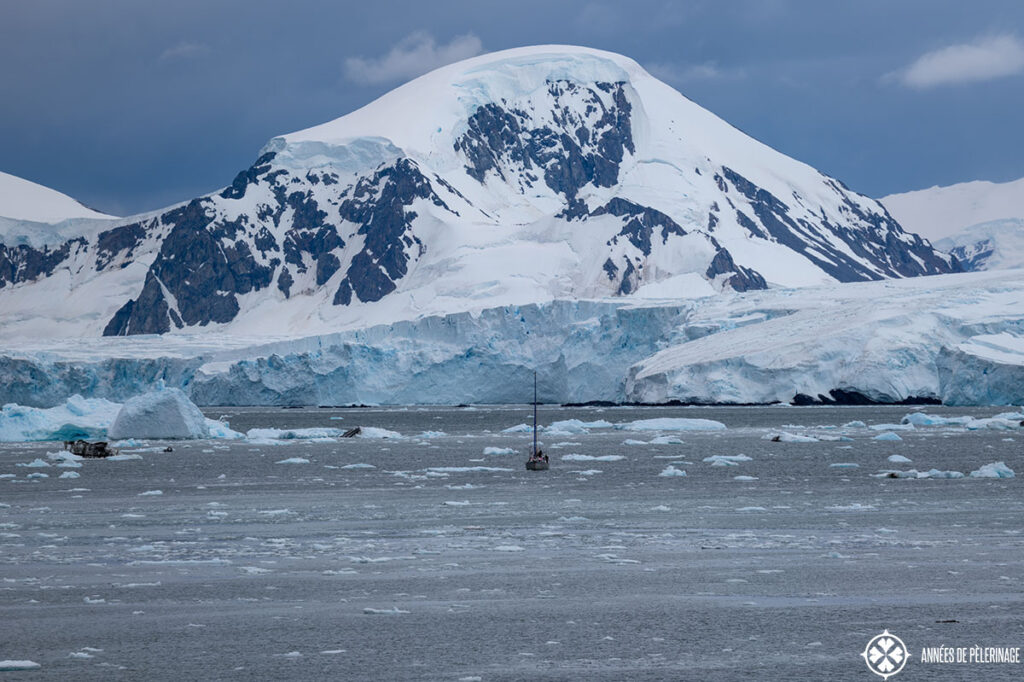 a tiny sail ship in a pack ice covered bay in antartica before a round snow-capped mountain